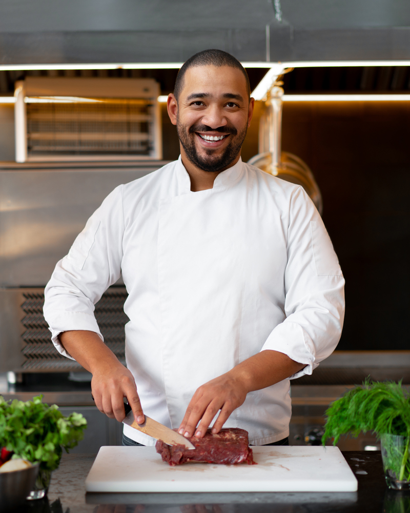 Handsome young African chef standing in professional kitchen in restaurant preparing a meal of meat and cheese vegetables